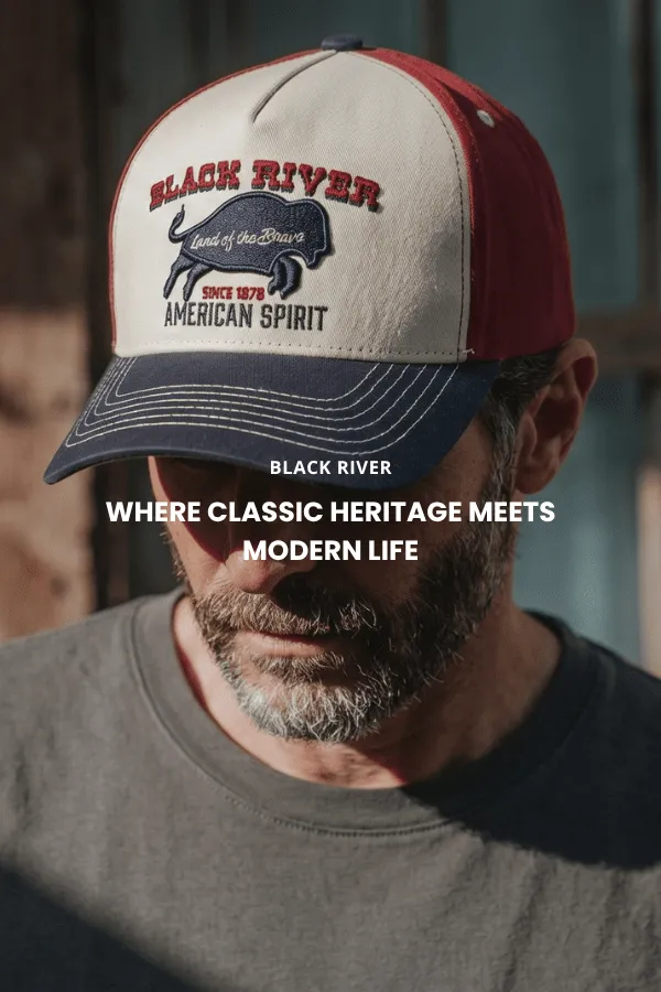 A man sits on a wooden crate in a studio wearing a classic brown felt cowboy hat with a wide brim, a dark denim shirt and jeans.