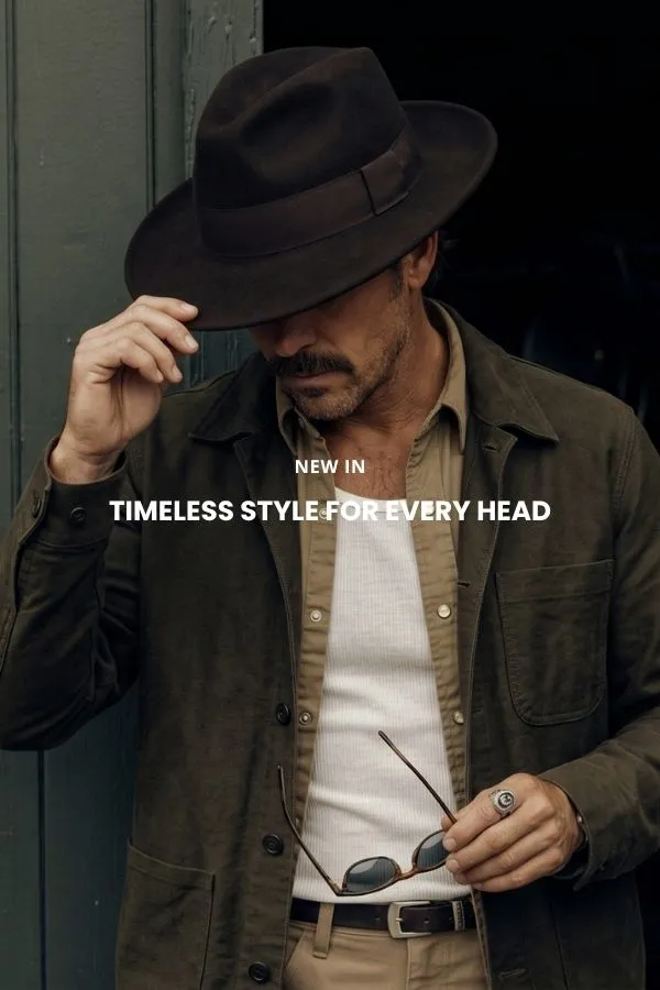 A man sits on a wooden crate in a studio wearing a classic brown felt cowboy hat with a wide brim, a dark denim shirt and jeans.